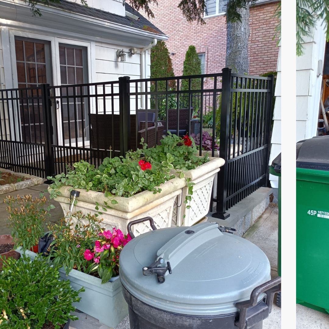 A home with white siding and dark trim has an outdoor patio area enclosed by a black metal fence, featuring potted plants with red and pink flowers, and a large, lidded gray trash can in the foreground.