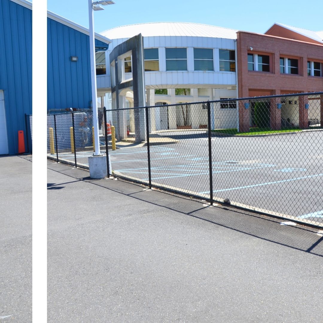 A paved parking lot with blue-painted parking lines is separated by a black chain-link fence from a large, modern building with a curved, light-colored facade and many windows, and a brick building on the right under a clear blue sky.