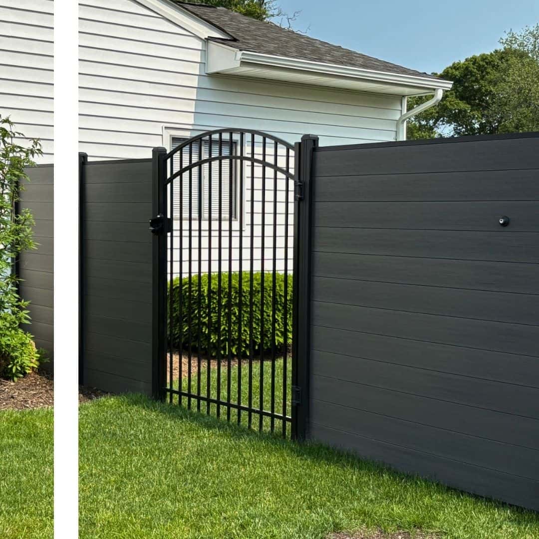 A modern dark grey horizontal slatted fence with a black arched metal gate stands on a manicured green lawn, with a white house in the background.
