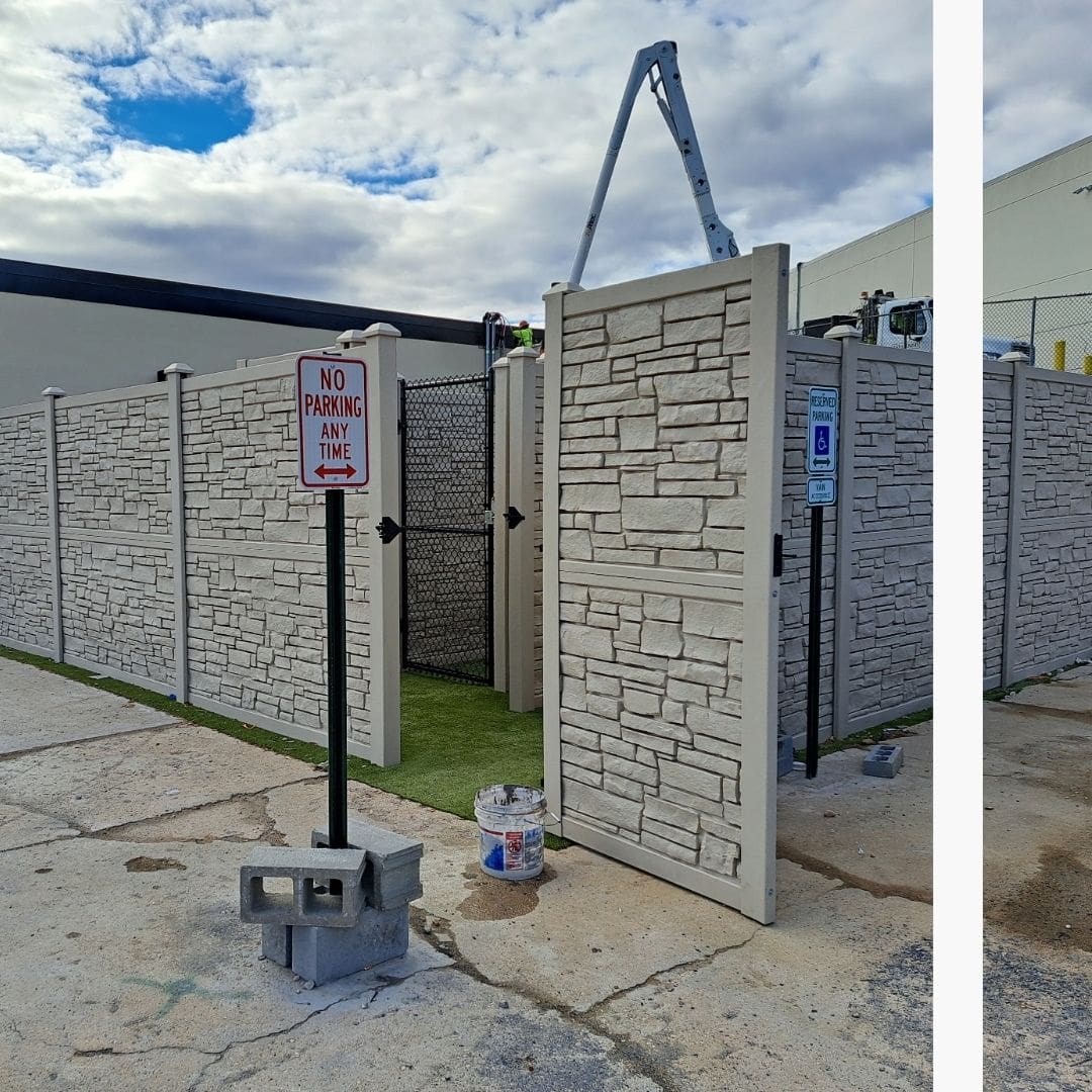 sturdy light-colored fence with a stone-like texture encloses an area with artificial grass, featuring a "No Parking Any Time" sign and a "Reserved Parking" sign, and a partial view of a lift in the background under a partly cloudy sky.