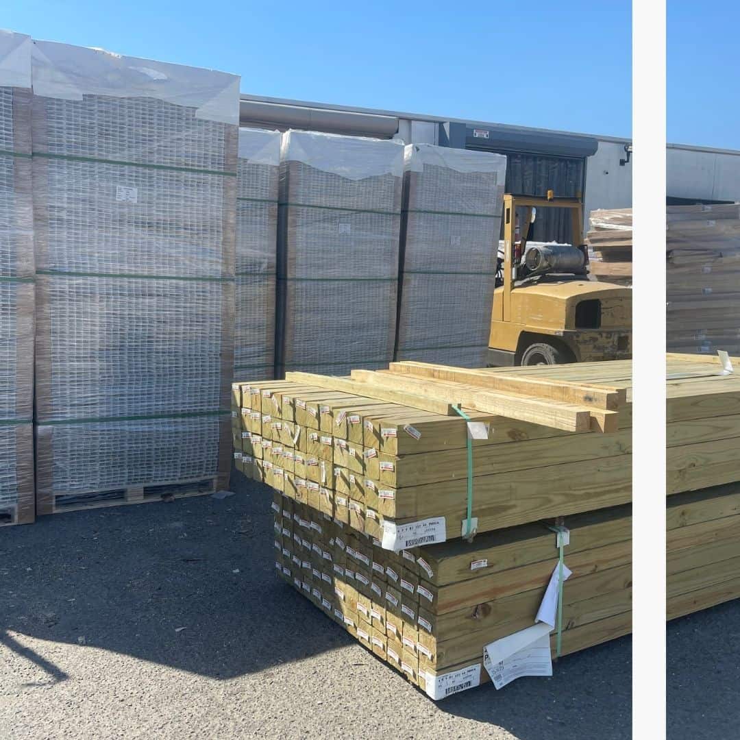 Stacks of pressure-treated lumber are neatly piled in an outdoor storage area, with shrink-wrapped pallets of fencing materials and a yellow forklift in the background near a building with loading docks, under a clear blue sky.