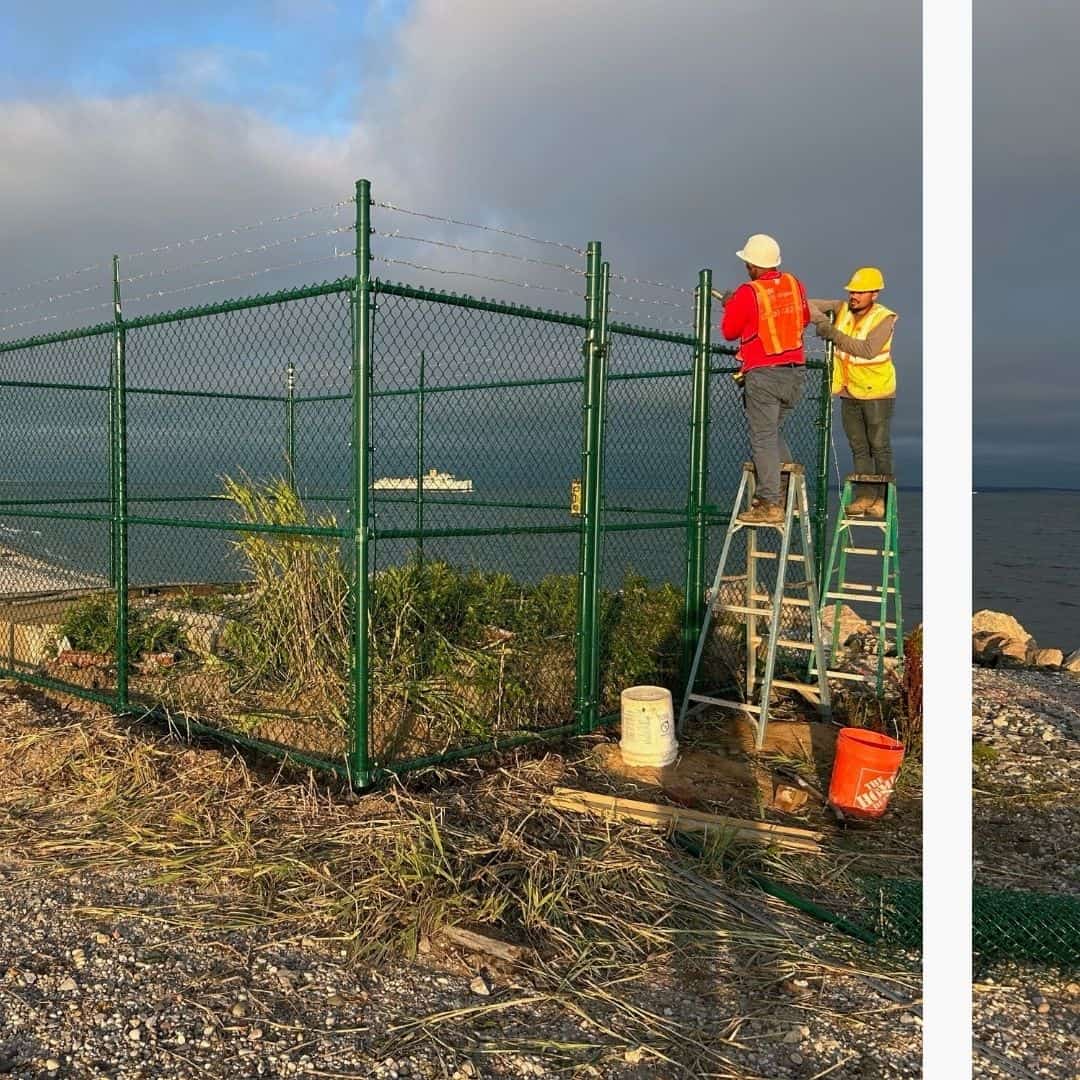 Two workers in safety gear install a green chain-link security fence with barbed wire next to the ocean.